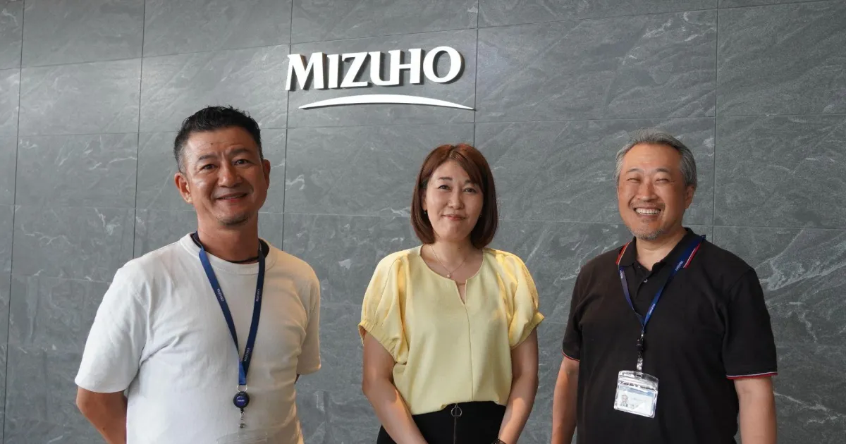 株式会社みずほ銀行ロゴの前に立つ外為営業部 総括チーム3名の写真